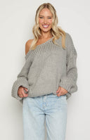blonde model wearing a grey oversized chunky knit sweater with an asymmetric neckline paired with white jeans