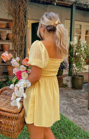 Holding flowers, the model is faced back to the camera, featuring a yellow mini dress. The dress has a speckled texture on the sleeves and lower half of the dress. The bust has a ruching fabric and is unlined.